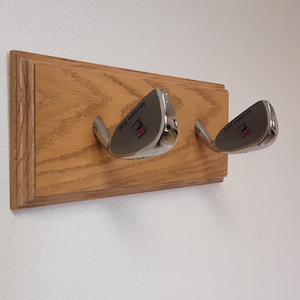 May include: A wooden wall-mounted rack with two silver golf club hooks. The hooks are shaped like golf clubs and have the text "Remarkable Model" printed on them.