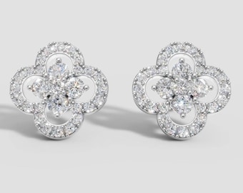 May include: A pair of silver-toned clover-shaped stud earrings. Each earring features a central cluster of round, clear gemstones surrounded by a halo of smaller, sparkling stones. The earrings are set against a neutral background.