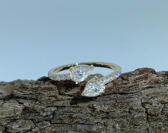 May include: A gold ring with two pear-shaped, clear gemstones. The ring band is encrusted with small, clear gemstones. The ring is displayed on a piece of textured, brown wood.