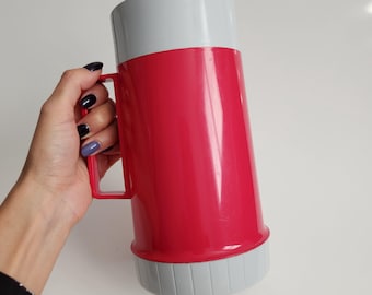 May include: A vintage red and gray thermos with a handle. The thermos has a red body, a gray top, and a gray base. The thermos is held by a hand with painted nails.