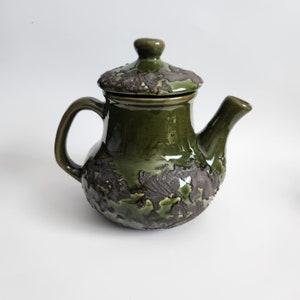 May include: A green ceramic teapot with a lid and a textured surface. The teapot has a brown glaze with a crackled effect.