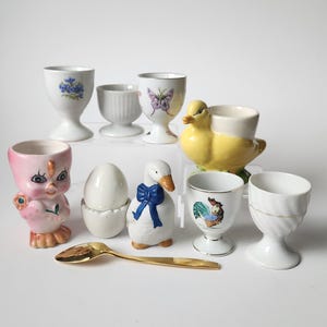 May include: A set of three ceramic mugs with a speckled, cream-coloured glaze. Each mug features a different hand-painted design: a blue and green leaf, a yellow flower, and a red berry. The mugs have a simple, modern design.