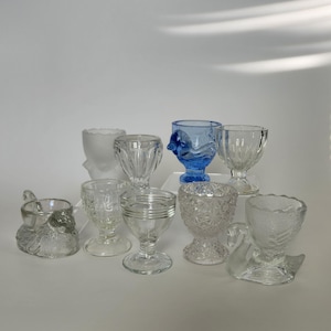 May include: A selection of clear and blue glass egg cups in diverse designs. The egg cups feature different shapes, including a swan, a bird, and fluted and patterned styles. Some are on pedestals.