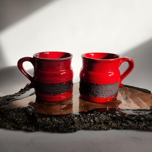 May include: Two red ceramic mugs with dark brown textured bands, each with a curved handle. The mugs sit on a dark wooden slab with a natural edge, against a white background.