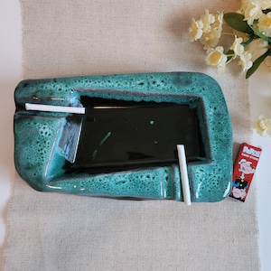May include: A teal ceramic ashtray with a dark green interior. Two white cigarettes are placed on the ashtray. A pack of Popeye Candy Sticks is next to the ashtray. The ashtray has a textured, speckled finish.