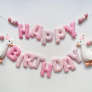 May include: Pink felt banner with the words "Happy Birthday" and two white felt geese with pink hats. The banner is strung with pink felt balls.