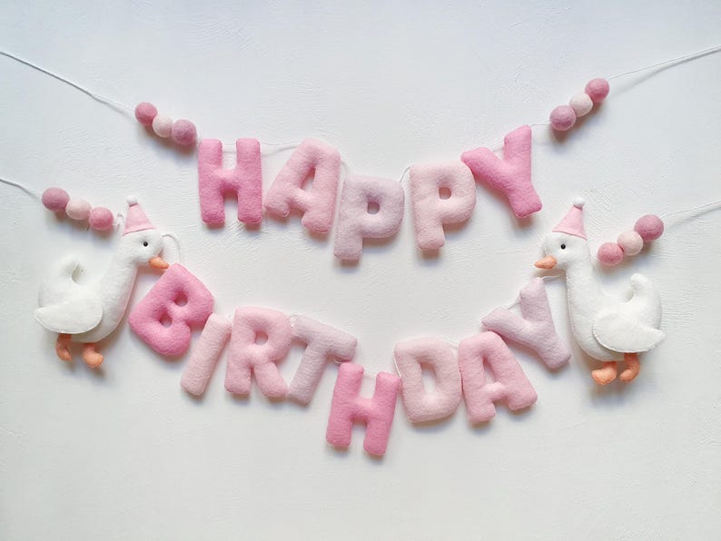 May include: Pink felt banner with the words "Happy Birthday" and two white felt geese with pink hats. The banner is strung with pink felt balls.