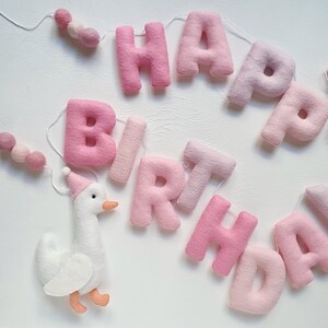 May include: Pink felt banner with the words "Happy Birthday" and a white felt goose with a pink hat. The banner is strung with pink pom-poms.