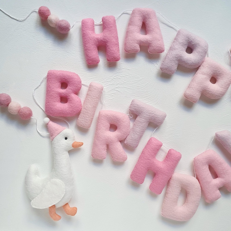 May include: Pink felt banner with the words "Happy Birthday" and a white felt goose with a pink hat. The banner is strung with pink pom-poms.