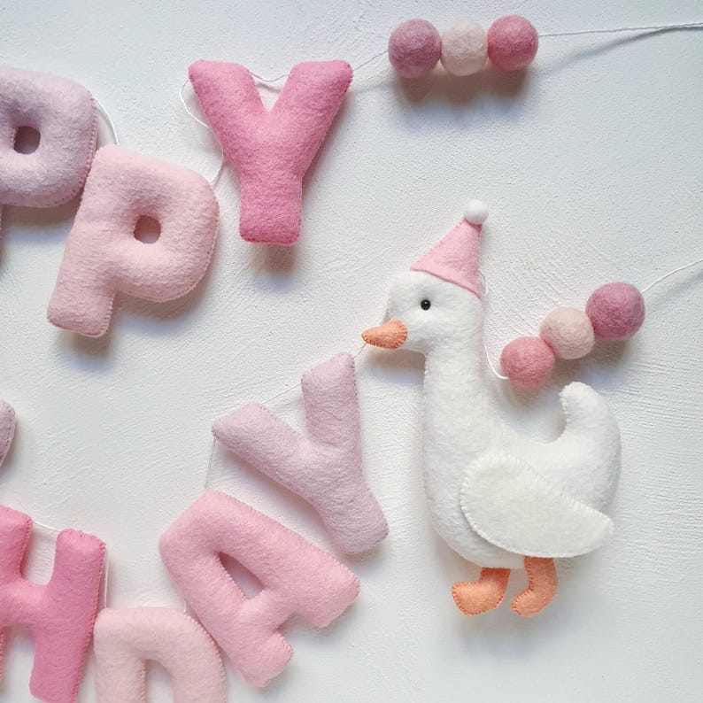 May include: Pink felt banner with the word "Happy" and a white felt goose wearing a pink party hat. The banner is strung with pink felt balls.
