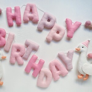 May include: Pink felt banner with the words "Happy Birthday" strung together with white thread. A white felt goose with orange feet is attached to the banner.