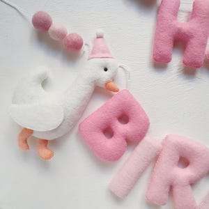 May include: A felt banner with the word "BIRTHDAY" spelled out in pink letters. A white felt goose with a pink hat is holding the letter "B".