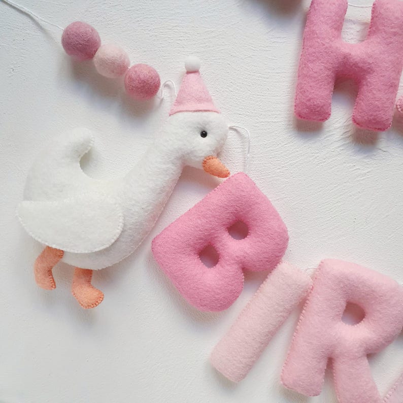 May include: A felt banner with the word "BIRTHDAY" spelled out in pink letters. A white felt goose with a pink hat is holding the letter "B".