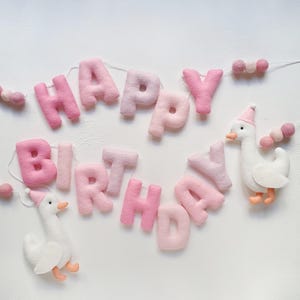 May include: A pink felt banner with the words "Happy Birthday" and two white felt geese with pink hats. The banner is strung with pink and white pom poms.