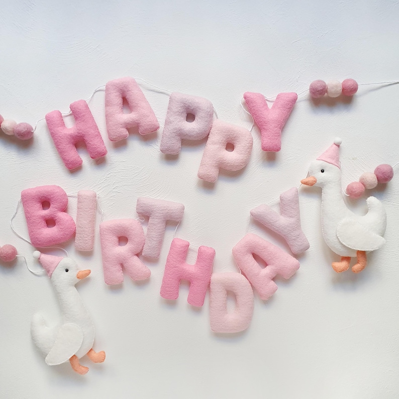 May include: A pink felt banner with the words "Happy Birthday" and two white felt geese with pink hats. The banner is strung with pink and white pom poms.