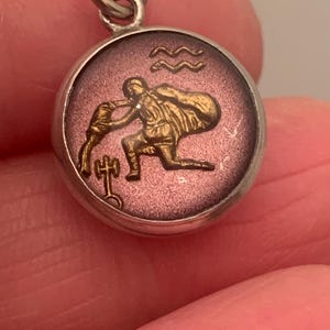 Puede incluir: Colgante redondo plateado con un relieve dorado detallado de una figura vertiendo agua, con el símbolo de Acuario. El colgante tiene un fondo morado y un lazo plateado para una cadena.