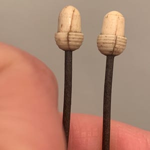 Könnte beinhalten: Zwei antike Hutnadeln. Jede Nadel hat einen cremefarbenen, eichelförmigen Kopf mit vertikalen und horizontalen geschnitzten Linien. Die Köpfe sitzen auf schlanken, dunkelbraunen Metallstielen. Der Hintergrund ist einfarbiges, neutrales Grau.