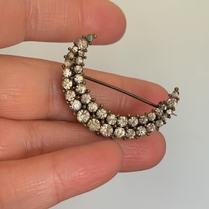 May include: A crescent moon brooch with clear, round rhinestones set in a gold-toned metal. The brooch has a pin on the back for secure fastening. The rhinestones are arranged in rows, creating a sparkling effect. A vintage-inspired jewelry piece.