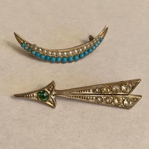May include: Two vintage brooches. One is a crescent moon shape with turquoise and pearl beads. The other is an arrow shape with a green gem at the center and clear gems along the arrow shaft. Both are gold-toned.