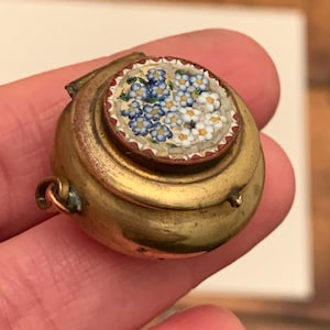 May include: A small, round, antique brass locket. The lid features a mosaic floral design with blue and white flowers and red and white accents. The locket has a hinged lid and a small loop for a chain.