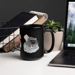 May include: A black ceramic mug with a white image of a chinchilla wearing sunglasses. The mug has a large handle and is sitting on a white surface.