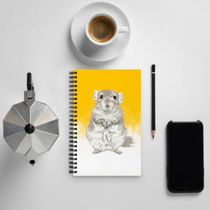 May include: A yellow spiral bound notebook with a white background and a cartoon illustration of a gray chinchilla. The chinchilla is sitting upright with its paws crossed and has a white fluffy tail.