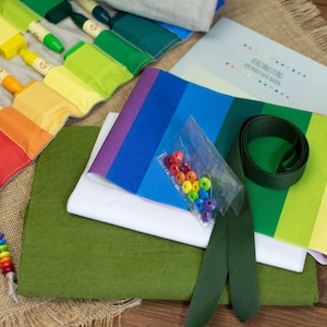 Könnte beinhalten: Ein farbenfrohes Lernset mit Regenbogen-Buntstiften, Stoff und Perlen. Das Set enthält einen Regenbogen-Buntstifthalter, grünen und weißen Stoff, ein Büchlein und eine Tüte mit bunten Perlen. Die Artikel sind auf einer Jutefläche angeordnet.