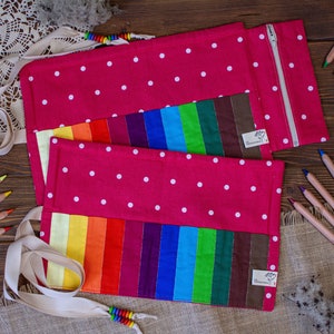 Könnte beinhalten: Eine rosa Stoff-Bleistiftrolle mit weißen Punkten und einem Regenbogen-Stofffutter. Die Rolle hat einen Reißverschluss und einen weißen Stoffgurt.