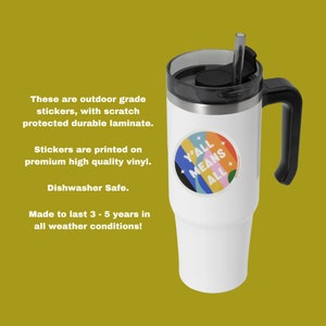 May include: White travel mug with a black handle and lid. The mug has a colorful sticker that says "Y'all Means All".
