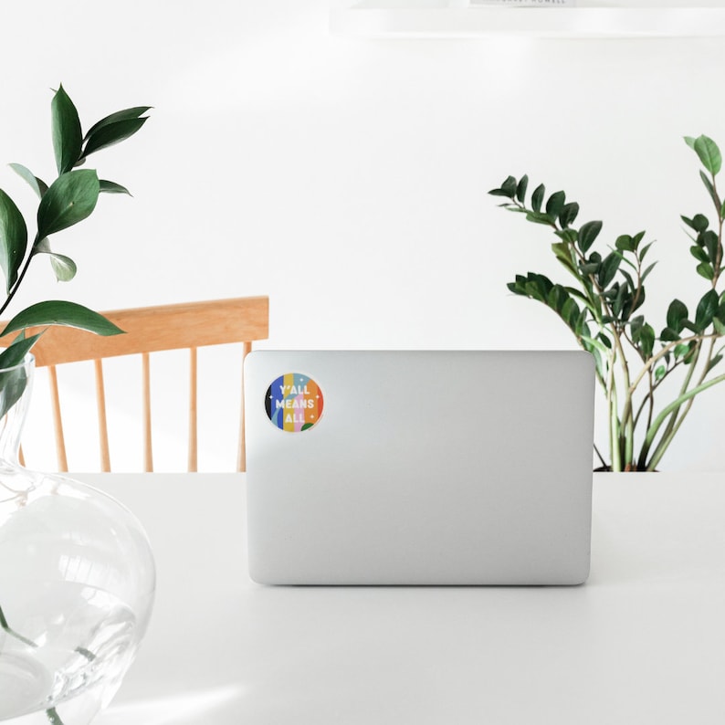 May include: A white laptop computer with a colorful round sticker that says "Y'all Means All" on the back.