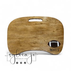 May include: A wooden lap desk with a football graphic and the name "MATT" in black. The desk has a handle and a curved design. The logo for "sylvia jo DESIGN CO" is in the lower left corner.