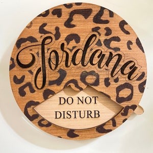 Puede incluir: Un colgador de puerta redondo de madera con un diseño de estampado de leopardo. El nombre "Jordana" está grabado en la mitad superior del colgador. La mitad inferior del colgador dice "No molestar".