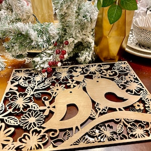 May include: A wooden laser cut placemat featuring a floral design and two birds perched on a branch. The placemat is brown and has a delicate, intricate design.