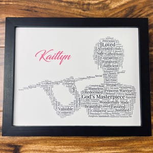 May include: A black and white word cloud art print in a black frame. The word cloud is shaped like a person and includes words like "loved", "forgiven", "redeemed", "God's Masterpiece", "valuable", "confident", and "chosen". The name "Kaittyn" is written in pink at the top of the print.