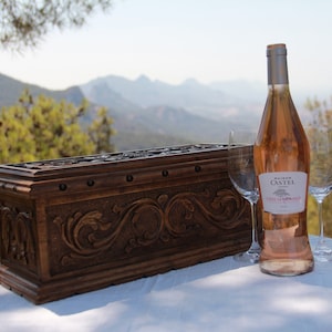 Puede incluir: Un cofre de madera tallada con adornos se encuentra sobre un mantel blanco, junto a una botella de vino rosado y dos copas de vino. La botella de vino tiene una tapa plateada y una etiqueta que dice "MAISON CASTEL CÔTES DE PROVENCE FRANCE". El fondo muestra un paisaje montañoso.