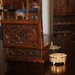 May include: A dark brown, intricately carved wooden jewelry box with an open lid, revealing a small compartment. A small, gold-colored trinket box with turquoise accents and a silver container sit nearby on a dark wooden surface. The scene suggests a vintage or antique theme.