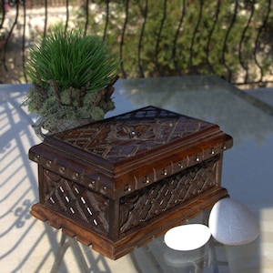 May include: A dark brown wooden box with intricate carved details, including geometric patterns and floral accents. The box is rectangular with a hinged lid and is displayed on a glass surface. Two white stones and a small plant are also visible.