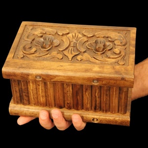 May include: A wooden box with intricate carvings on the lid. The box is brown and has a rectangular shape. It is being held in a person's hand.