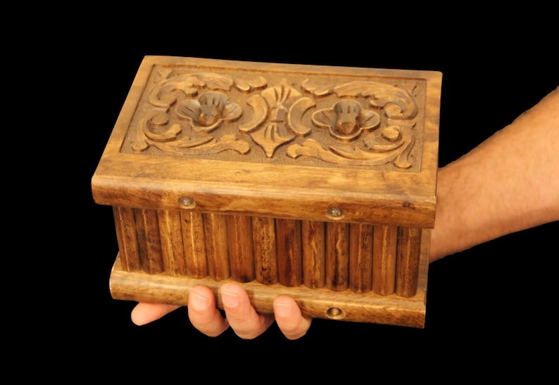May include: A wooden box with intricate carvings on the lid. The box is brown and has a rectangular shape. It is being held in a person's hand.
