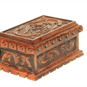 May include: A rectangular wooden box with intricate floral and scroll carvings. The box is stained a warm brown color and features decorative detailing around the edges. The lid and sides are adorned with carved designs.