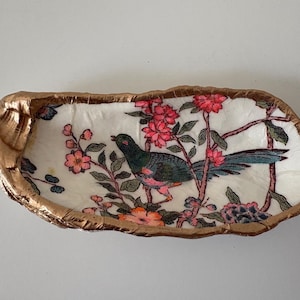 May include: An oval-shaped oyster shell dish with a metallic gold painted rim. The interior features a decoupage design of a dark green bird perched on branches with pink and orange flowers against a white background, creating a decorative tray.