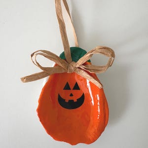 May include: A decorative oyster shell painted bright orange with a black jack-o'-lantern face featuring triangular eyes and a curved mouth. The shell has a green painted top and is adorned with a tan raffia ribbon tied in a bow at the top.