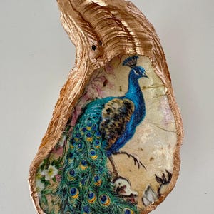 May include: A decorative oyster shell with a metallic gold exterior. The interior features a printed image of a blue peacock with its tail feathers fanned out. Floral accents are also visible.