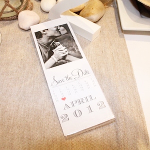 May include: A white card with a black and white photo of a couple kissing. The card says "Save the Date" and has a calendar with a red heart marking April 2012.
