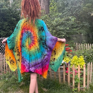 Op de afbeelding: Een levendige tie-dye kimono met een spiraalpatroon, met een spectrum aan kleuren waaronder oranje, geel, groen, blauw en paars. De lichte stof valt over de schouders en creëert een vloeiend silhouet. Het kledingstuk wordt buiten gedragen.