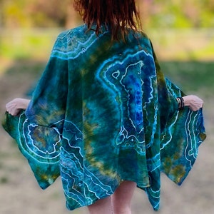 May include: A tie-dye kimono in shades of teal, blue, and gold, with a white outline pattern. The garment has wide sleeves and a flowing silhouette, suggesting a bohemian or artistic style. The person is wearing the kimono.