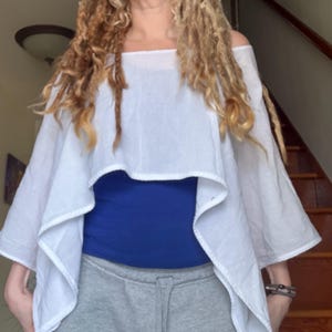 May include: White off-the-shoulder top with a layered design. The top is paired with a blue top and gray sweatpants. The top has a square neckline and a flowing, asymmetrical hemline.