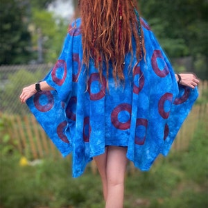Puede incluir: Una parte superior estilo kimono azul estampada con grandes diseños circulares de color rojo oscuro. La parte superior tiene mangas anchas y una silueta fluida. La tela parece tener un efecto tie-dye. La persona lleva la parte superior al aire libre.