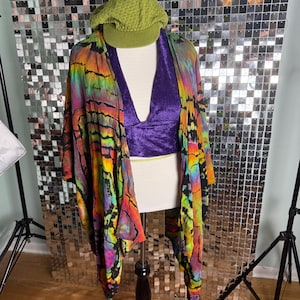 Op de afbeelding: Een mannequin toont een kleurrijke outfit. Het omvat een sjaal met tie-dye patroon, een dieppaarse fluwelen haltertop en een groene gebreide pet. De sjaal heeft levendige tinten rood, oranje, geel, groen en paars.