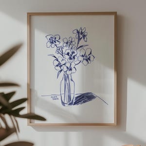 Pode incluir: Um desenho a tinta azul emoldurado de flores num vaso está pendurado acima de uma gira-discos laranja com um disco na plataforma giratória. Um álbum de vinil com um gráfico de banana amarela está à esquerda.